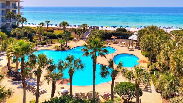 A resort with a large pool, palm trees, and a beach with umbrellas.