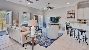 A spacious living area with a sofa, coffee table, and a TV. There are ceiling fans, a kitchen area with bar stools, and a dining table.