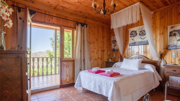 Ein Zimmer mit Holzboden, einem Bett, einem Nachttisch, einem Sessel, einem Fenster mit Vorhängen und einem Balkon.
