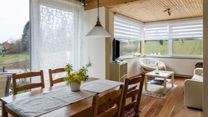 Uma sala de estar aconchegante com uma mesa de jantar, cadeiras, um vaso de planta, uma TV e uma janela com persianas.