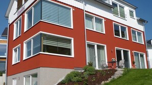Uma casa de dois andares, em vermelho e branco, com varanda frontal, um pequeno jardim e escadas que levam à entrada.