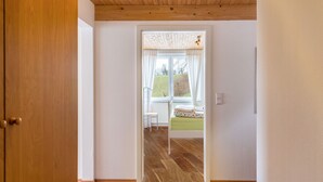 Quarto com piso de madeira, cama com colcha verde e janela com cortinas brancas, com vista para uma paisagem verde.