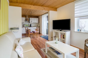 Un salon moderne avec un canapé blanc, une table basse, un téléviseur à écran plat et un coin repas avec des chaises en bois, et une cuisine en arrière-plan.