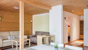 Ein gemütliches Zimmer mit Holzbalkendecke, einer Couch, einem kleinen Tisch, einem Bücherregal und einem Bett mit weißer Bettdecke.