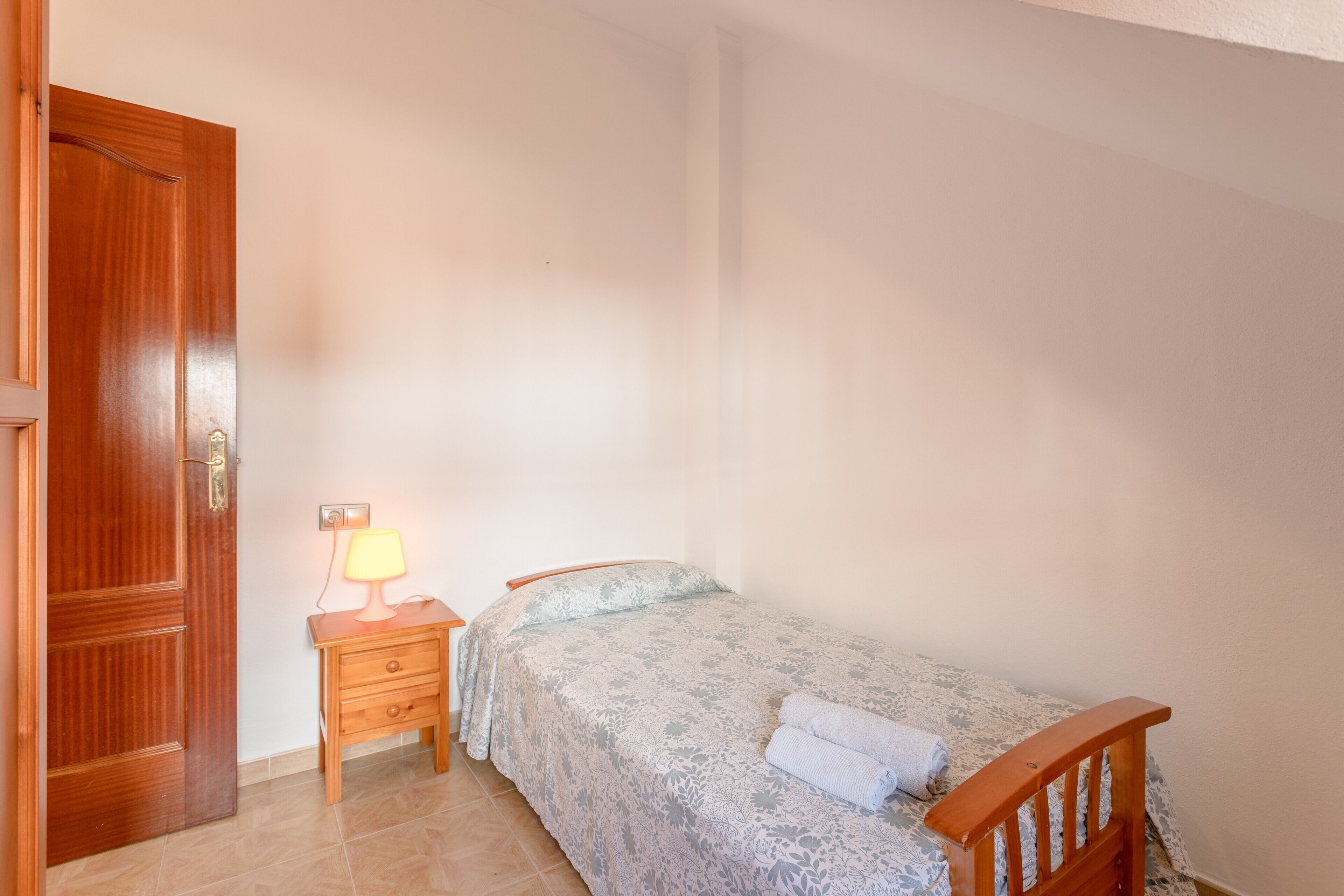 A bedroom with a bed, a nightstand with a lamp, a wooden door, and a tiled floor.