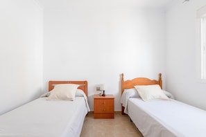 A small, clean room with two single beds, a wooden nightstand with a lamp, and a window.