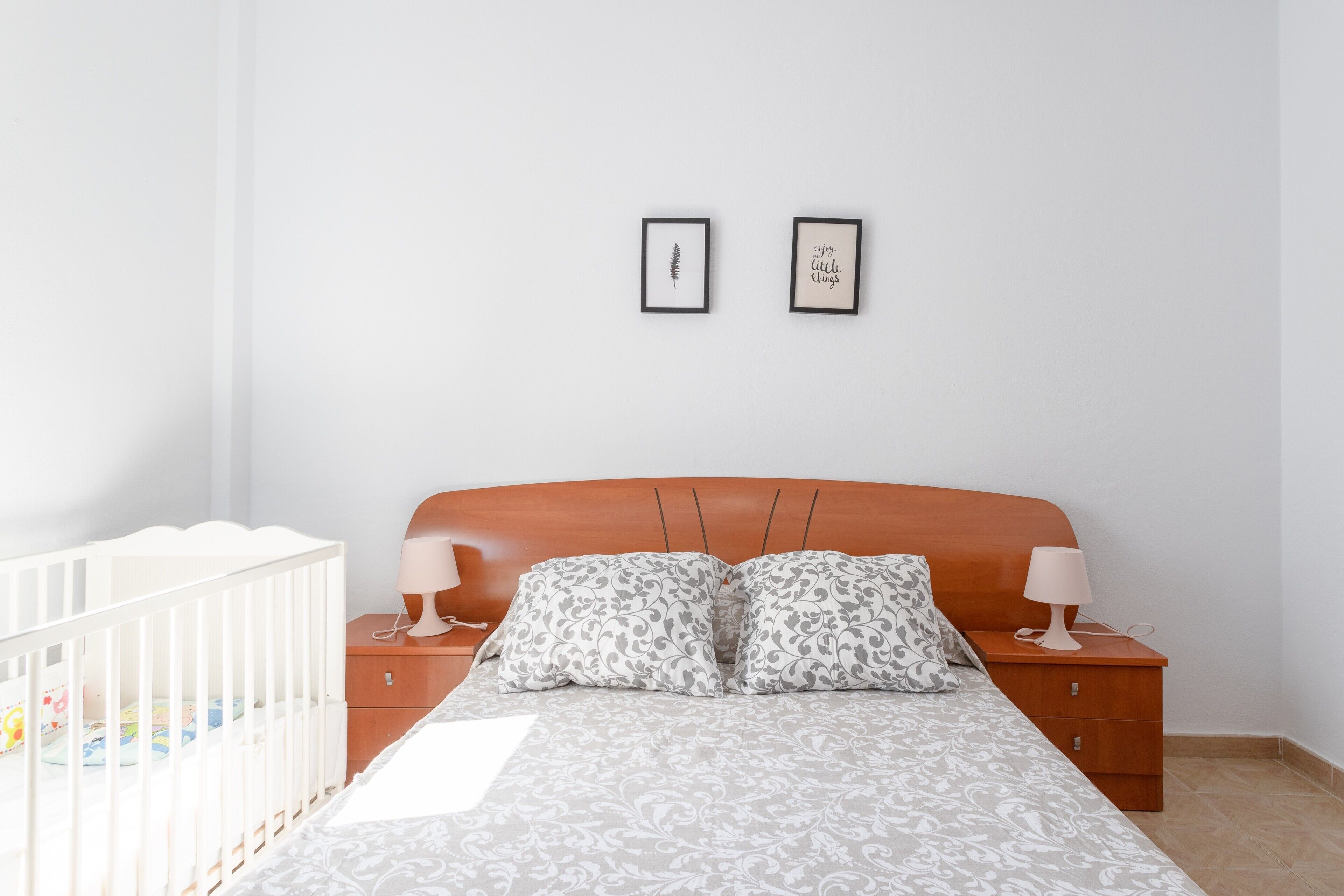 A bedroom with a wooden headboard, two bedside tables with lamps, a crib, and two framed pictures on the wall.