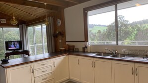 A kitchen with wooden cabinets, a sink, a window with a view of greenery, a clock, and a television on a wooden stand.