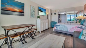 Een kamer met een houten eettafel, een bed met een kleurrijk beddegoed, een witte dressoir en uitzicht op de oceaan door een schuifdeur.
