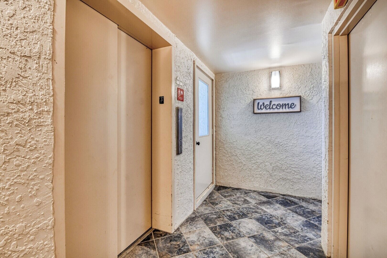 Ein Flur mit einem 'Welcome'-Schild, einer Tür mit Nummer und einer strukturierten Wand.