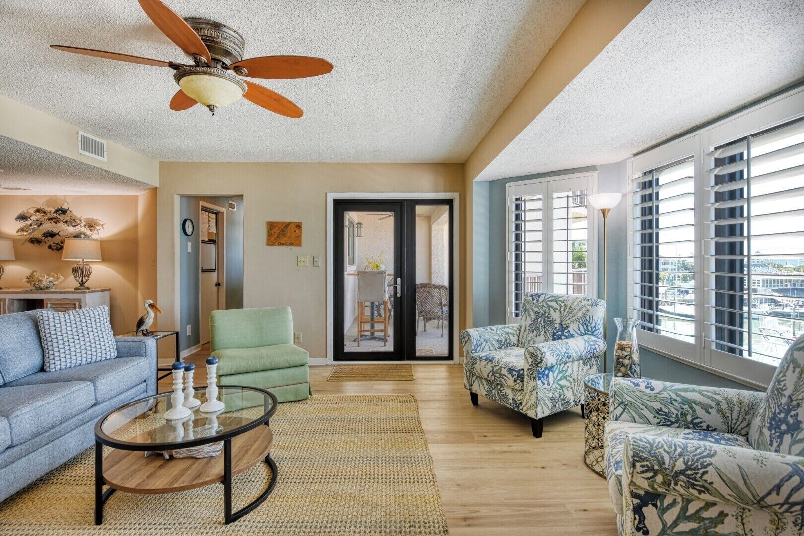 A spacious living room with a ceiling fan, a sofa, two armchairs, a coffee table, and a view through a glass door to an outdoor area.