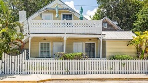 Une maison à deux étages avec un balcon, une clôture en bois peinte en blanc et des plantes tropicales.