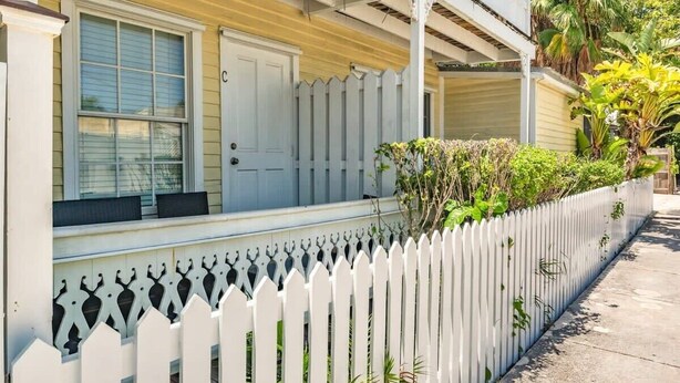 Une maison à deux étages avec une clôture en bois peinte en blanc, une véranda et un petit jardin agrémenté de plantes et d’un palmier.