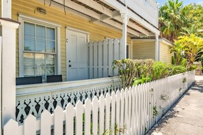 A two-story house with a white picket fence, a covered porch, and a small garden area with plants and a palm tree.