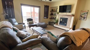 Ein Wohnzimmer mit einem Ecksofa, einem Couchtisch und einem Kamin. Ein Fernseher ist an der Wand montiert und ein Deckenventilator vorhanden.