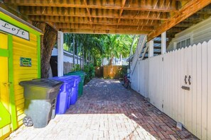 Un étroit sentier pavé de briques mène à une zone abritée, dotée d’un plafond en bois et d’une clôture blanche en piquets.