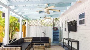 Un espace extérieur abrité, aménagé avec des meubles en osier, un ventilateur de plafond et un téléviseur fixé au mur.