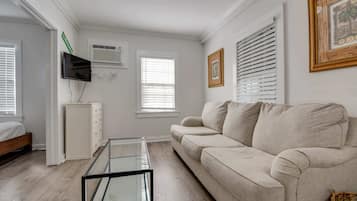 A living room with a sofa, a glass coffee table, a TV mounted on the wall, and a bed with a wooden headboard.