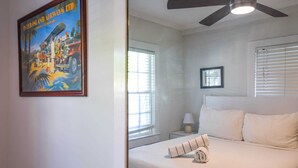 Une chambre d’hôtel avec un lit, un ventilateur de plafond, une fenêtre avec des stores et un tableau encadré au mur.