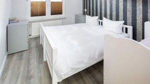 A bedroom with a white bed, a radiator, a window with blinds, a bedside table, and a striped wallpaper.