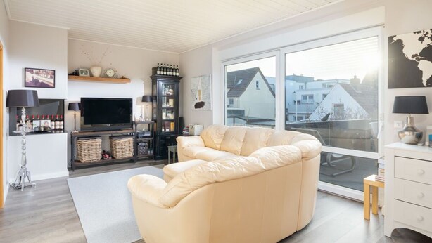 A modern living room with a beige sofa, a flat-screen TV, a wine rack, and a large window offering a view of buildings.