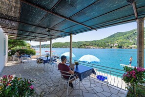 A covered outdoor seating area with tables and chairs, overlooking a body of water and a hillside.