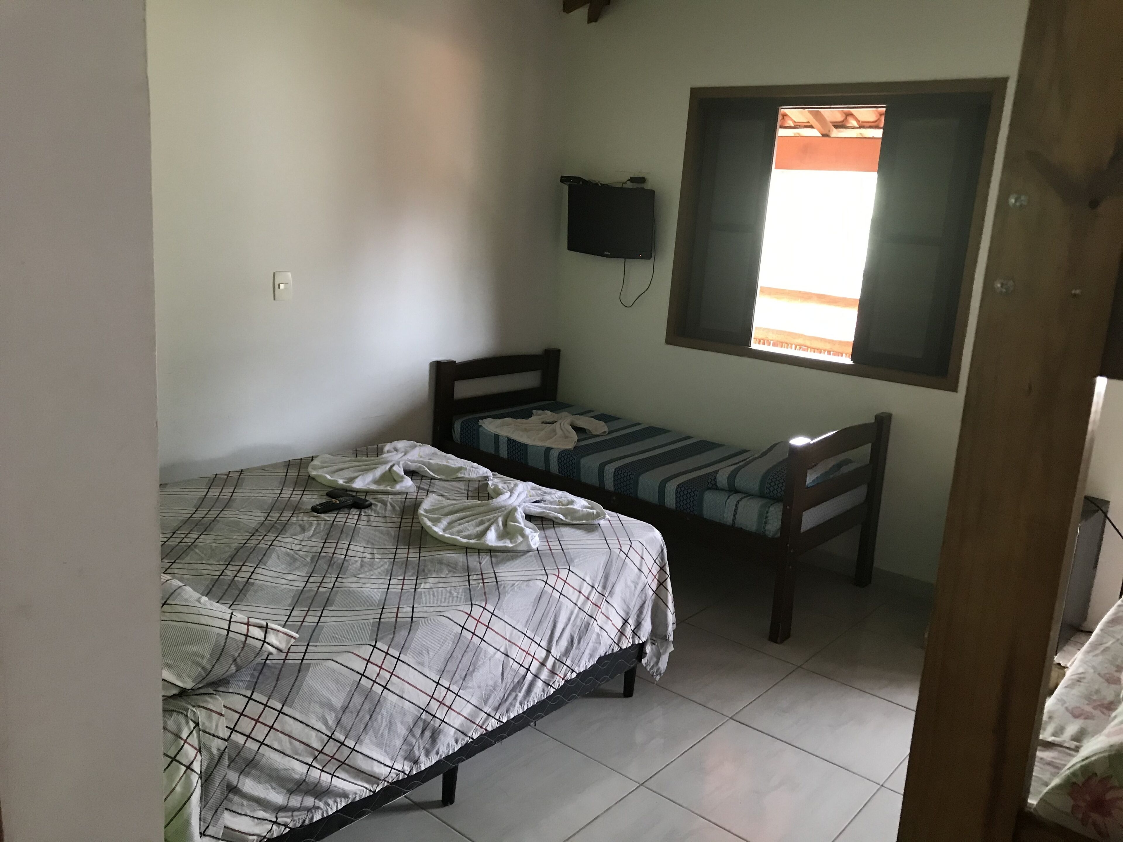 A small, sparsely furnished room with a bed, a bench, a television mounted on the wall, and a window.