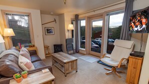 A cozy living room with a brown leather sofa, a wooden coffee table, and a white armchair. There is a fireplace, a TV mounted on the wall, and a large window offering a view of parked cars and trees. - Tyra Lookout Condo: Ski-In/Ski-Out (Breckenridge)