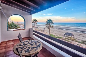 Een balkon met een geruite tafel en stoelen, uitkijkend op een strand met palmbomen en een rieten hut.