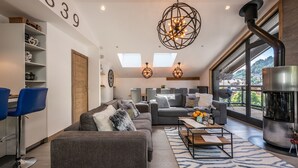 A modern living room with a grey sofa, blue chairs, a coffee table, and a fireplace. There are skylights and a large window offering a view of the outdoors. - OZIGO - Modern Apartment in Luxury Residence (Les Gets)