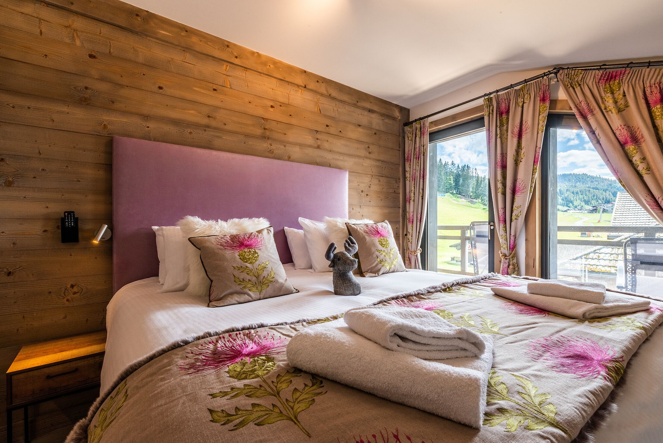 A bedroom with a wooden headboard, a large bed with floral bedding, a nightstand, and a view of a green landscape through the window.