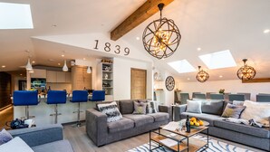 A modern living room with a grey sofa, blue bar stools, a coffee table, and a large window with skylights. - OZIGO - Modern Apartment in Luxury Residence (Les Gets)
