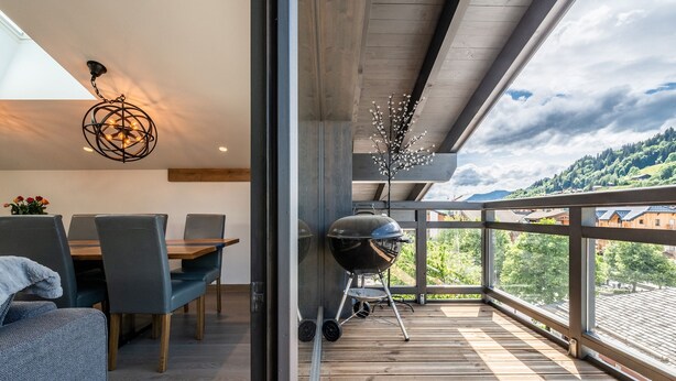A modern dining area with a wooden table and chairs, a grill, and a view of a mountainous landscape through large windows. - OZIGO - Modern Apartment in Luxury Residence (Les Gets)
