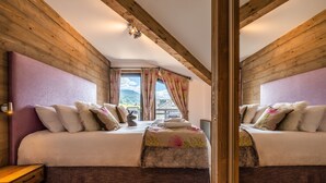 A bedroom with two beds, wooden beams, a window with curtains, and a view of the mountains. - OZIGO - Modern Apartment in Luxury Residence (Les Gets)