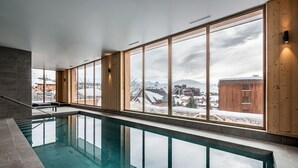 A modern indoor swimming pool with large windows offering views of a snowy landscape and buildings. - FAGUS - Spacious Apartment in Luxury Residence (Huez)