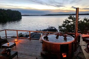 A wooden deck with a hot tub, outdoor seating, and a view of a lake at sunset.