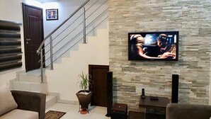 A modern living room with a staircase, a flat-screen TV mounted on a stone wall, and a potted plant. - Esenyo Luxury Apartments (Abuja)