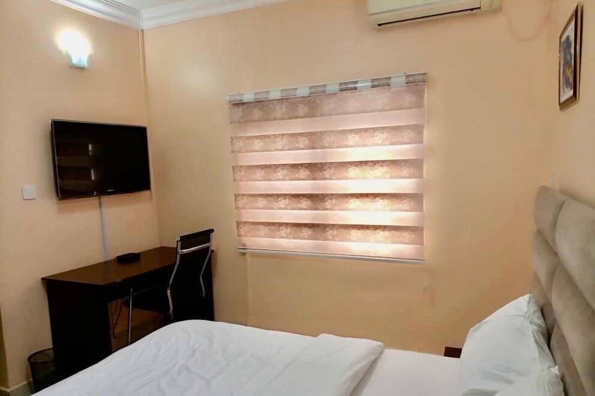 A bedroom with a bed, a desk, a chair, a TV, and a window with blinds.