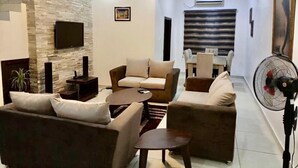 A modern living room with a stone accent wall, a flat-screen TV, and a dining area with a table and chairs. - Esenyo Luxury Apartments (Abuja)