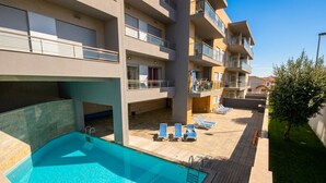A swimming pool with lounge chairs, a wooden deck, and a multi-story building with balconies. - Baia Residence 2 - Prata By SCH (São Martinho do Porto)