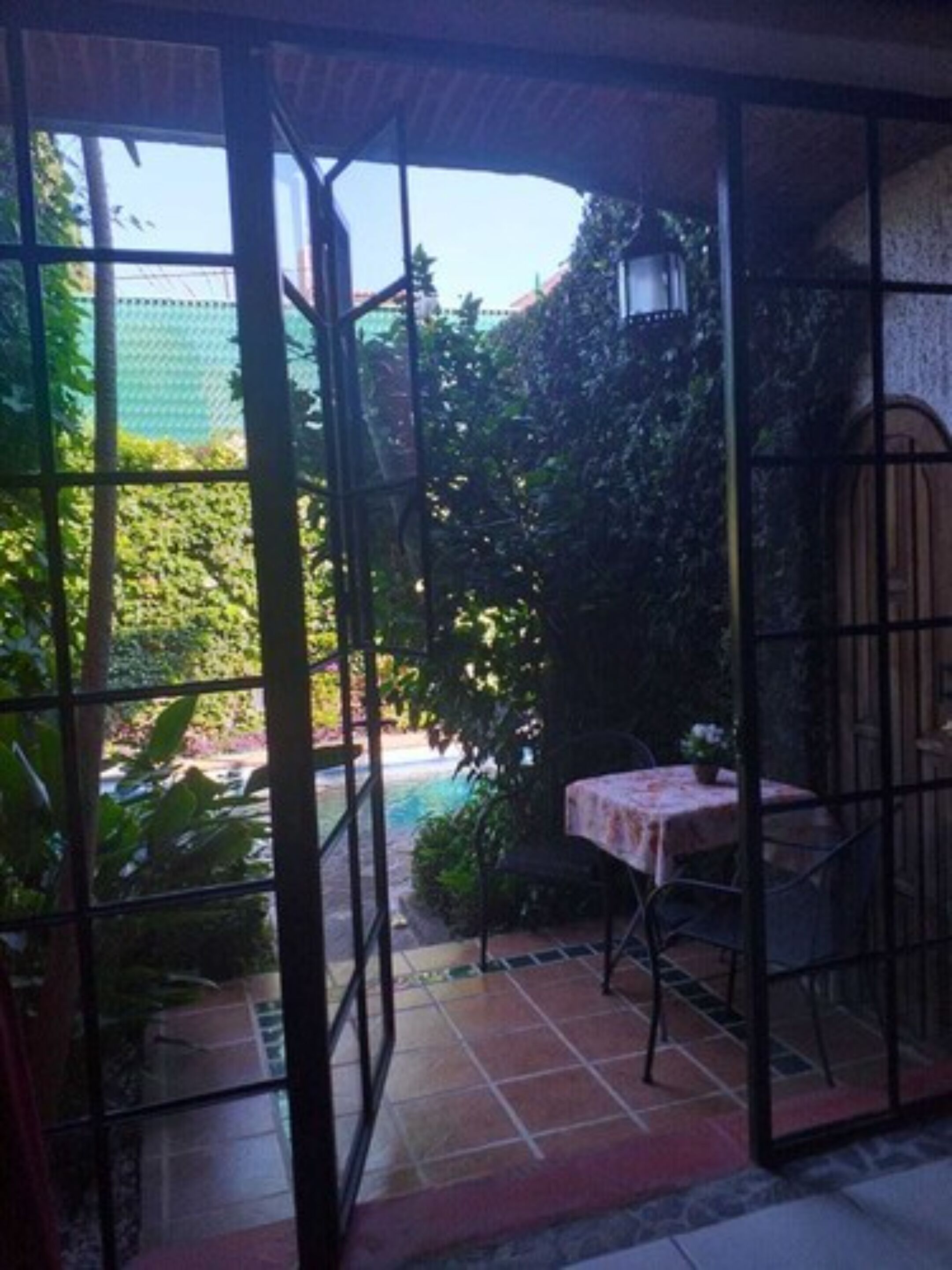 Un patio cubierto con mesa y sillas, rodeado de follaje y vistas a una piscina.