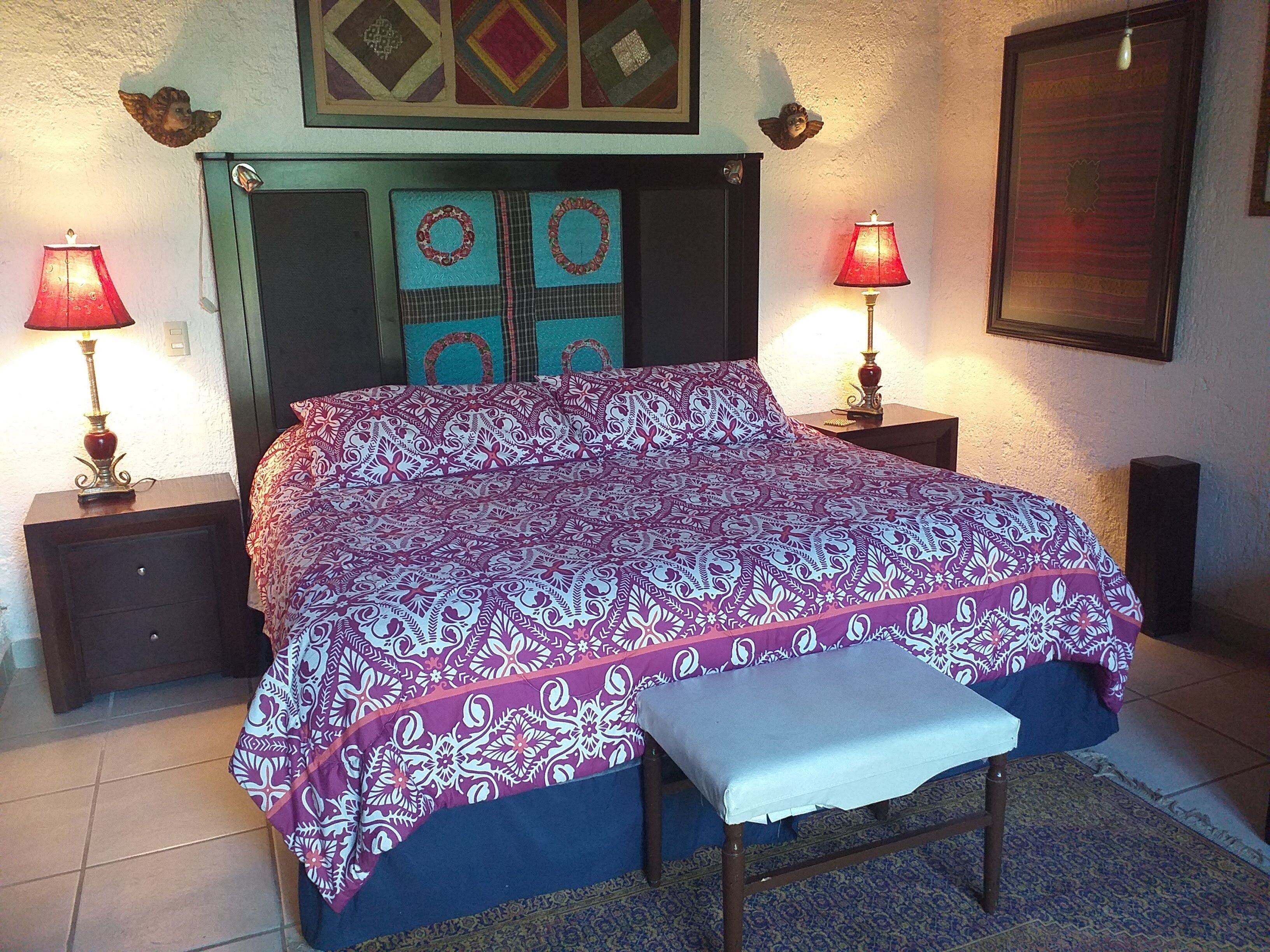 Una camera da letto con una coperta a motivi, due lampade da comodino, una panca e decorazioni murali.