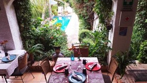An outdoor patio with a dining table set for two, surrounded by chairs, with a pool and greenery in the background. - Casa Flores Bed & Breakfast (Ajijic)