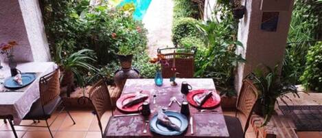 Un patio al aire libre con una mesa de comedor dispuesta para dos personas, rodeada de sillas, y una piscina y follaje al fondo.