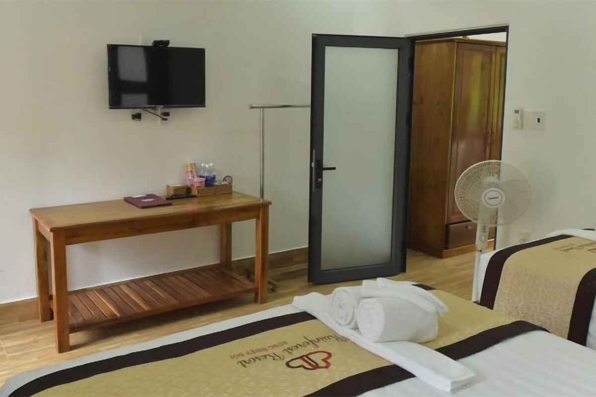A hotel room with a bed, a bedside table with toiletries, a TV mounted on the wall, a door, and a fan.