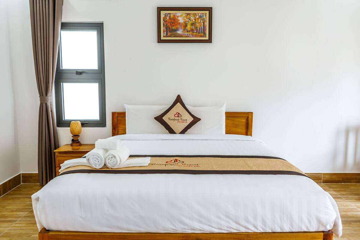 A neatly made bed with a white comforter and a brown decorative pillow, a wooden nightstand with a lamp, and a window with curtains.