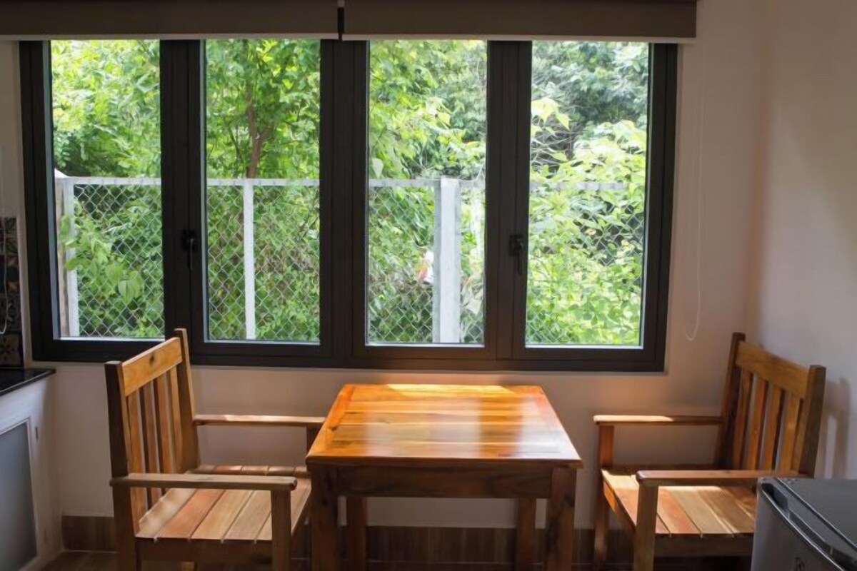 A room with a wooden table and two chairs, a window with a view of greenery, and a white wall.