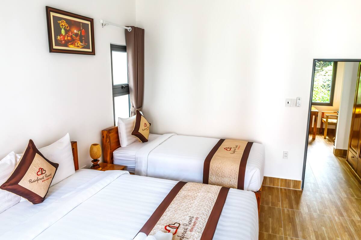 A hotel room with two beds, a painting on the wall, a window with curtains, and a view of the outside.