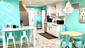 A modern kitchen with white cabinets, a dining area with a wooden table and turquoise chairs, and a colorful rug on the floor.
