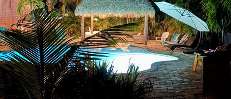 A nighttime scene with a swimming pool, a thatched-roof structure, and lounge chairs.